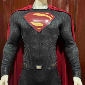 May include: A Superman costume featuring a black and gray textured bodysuit with a red and yellow emblem on the chest. The costume includes a red cape and is displayed on a mannequin. The suit has gold accents and is designed to resemble the superhero's iconic look.