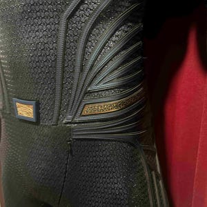 May include: Close-up of a dark blue costume with intricate detailing. The suit features a textured pattern and metallic accents in gold and blue. The design includes layered elements and a belt buckle.