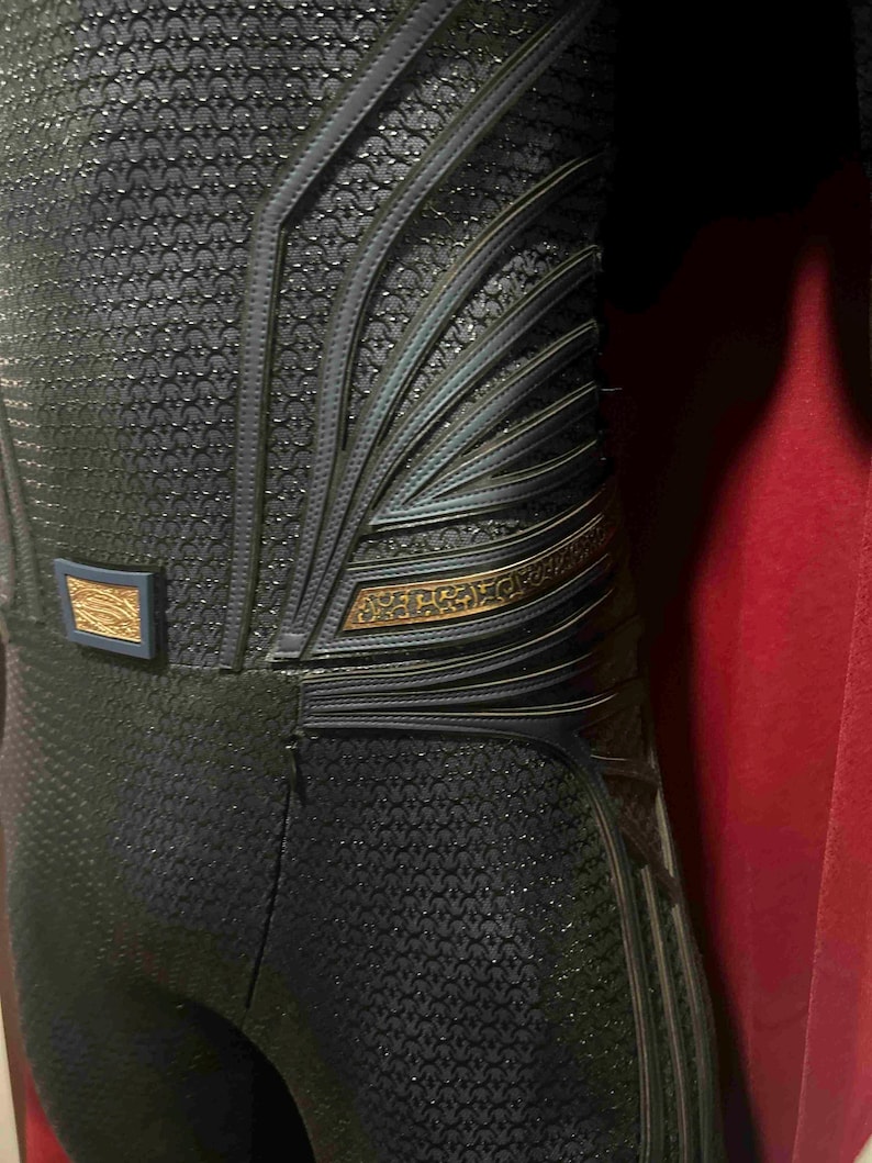 May include: Close-up of a dark blue costume with intricate detailing. The suit features a textured pattern and metallic accents in gold and blue. The design includes layered elements and a belt buckle.