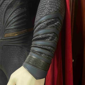May include: Close-up of a superhero costume featuring a textured, dark blue fabric with intricate detailing. The costume includes blue and black accents on the arm, with a red cape visible in the background. The arm is on a mannequin.