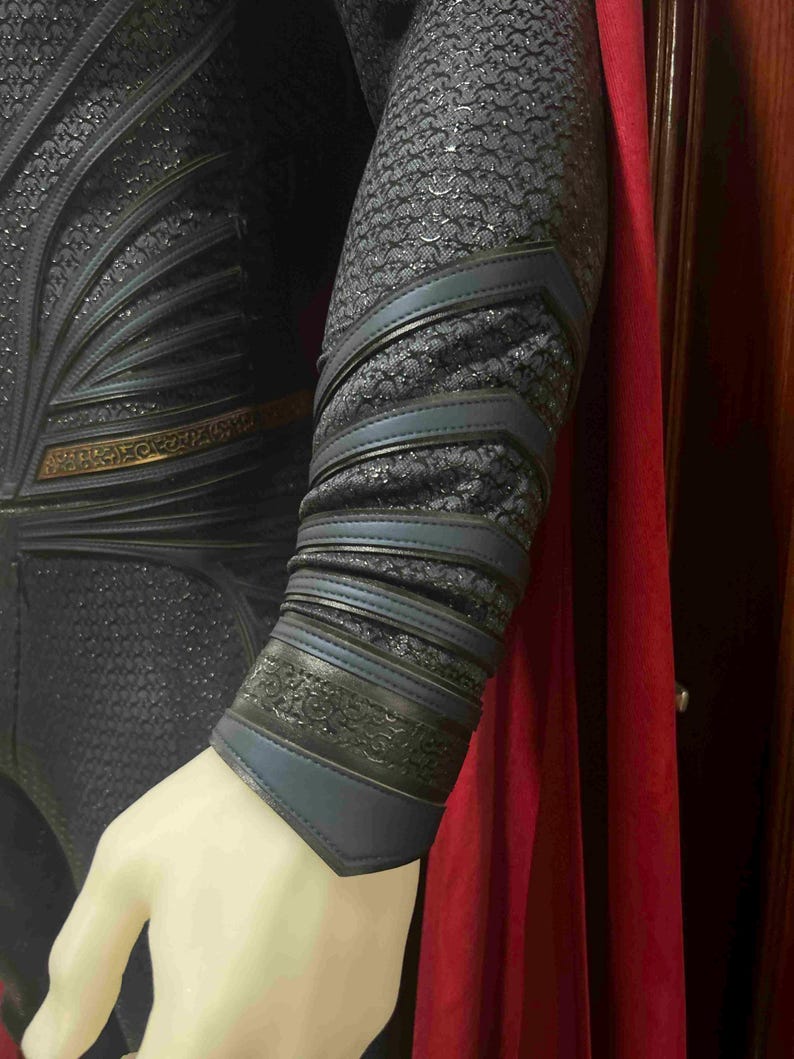 May include: Close-up of a superhero costume featuring a textured, dark blue fabric with intricate detailing. The costume includes blue and black accents on the arm, with a red cape visible in the background. The arm is on a mannequin.