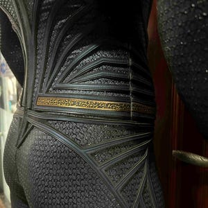 May include: Close-up of a black textured costume with intricate geometric designs. The costume features a gold-colored belt and detailed patterns, suggesting a superhero or fantasy theme. The fabric has a subtle sheen.