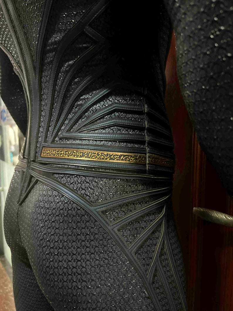 May include: Close-up of a black textured costume with intricate geometric designs. The costume features a gold-colored belt and detailed patterns, suggesting a superhero or fantasy theme. The fabric has a subtle sheen.