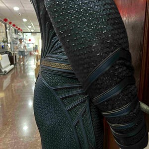 May include: A black and dark teal costume with a textured, scale-like pattern. The costume features geometric designs and dark teal accents, with a gold-colored belt. The sleeve has dark teal bands.