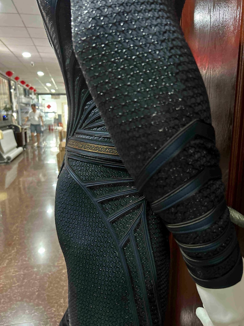 May include: A black and dark teal costume with a textured, scale-like pattern. The costume features geometric designs and dark teal accents, with a gold-colored belt. The sleeve has dark teal bands.
