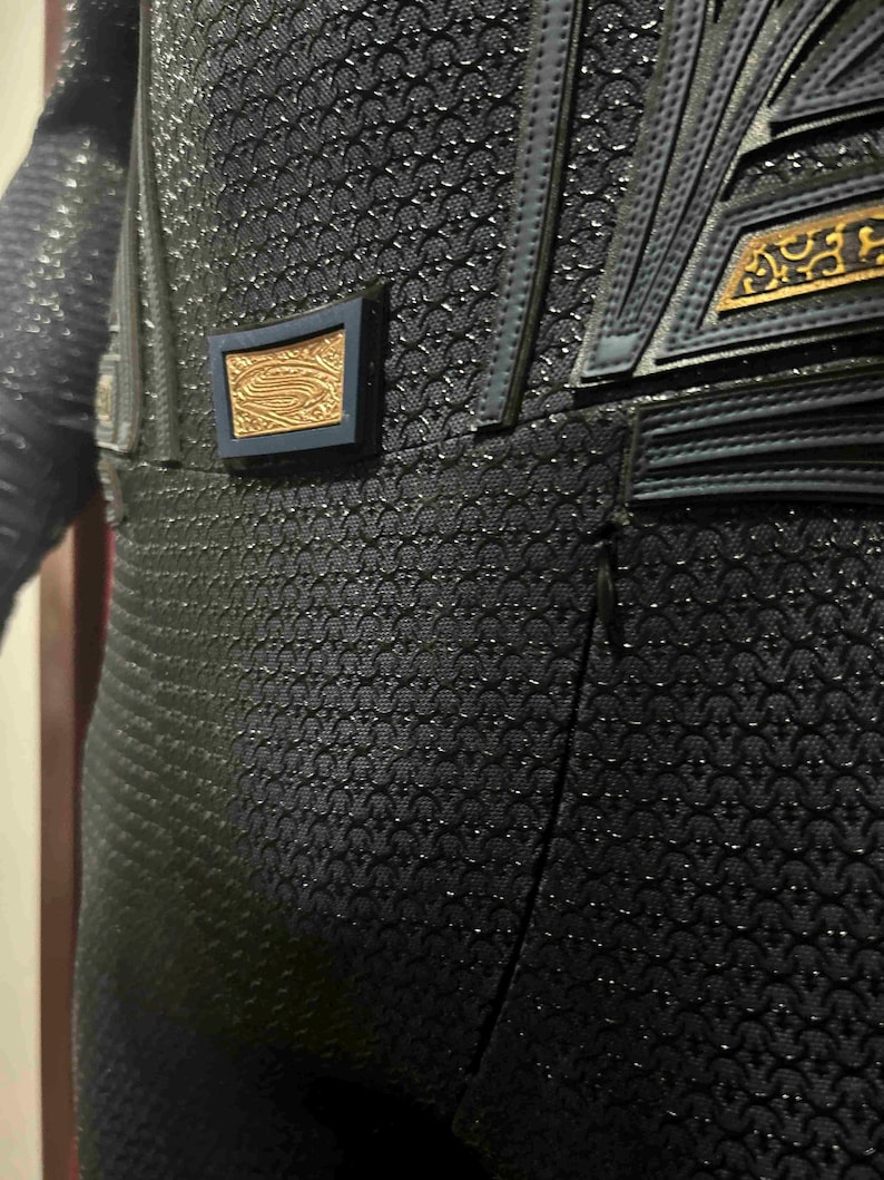 May include: Close-up of a black costume with a textured, scale-like pattern. The costume features dark blue accents and gold-colored decorative elements. A small, rectangular emblem with a gold design is visible.