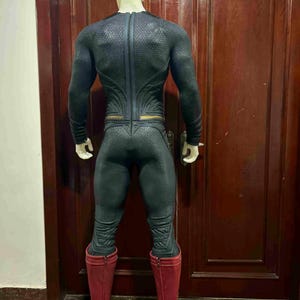 May include: A black and gray superhero costume with a textured pattern, featuring a full-body design with long sleeves and a high collar. The costume includes red boots and gold accents. The mannequin is positioned in front of a dark brown door.