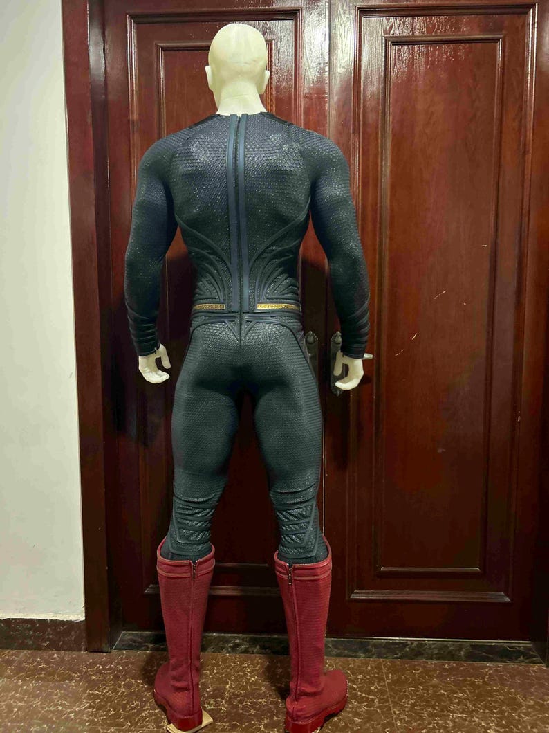 May include: A black and gray superhero costume with a textured pattern, featuring a full-body design with long sleeves and a high collar. The costume includes red boots and gold accents. The mannequin is positioned in front of a dark brown door.