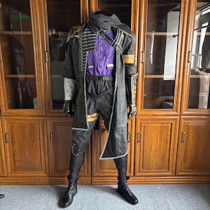 Könnte beinhalten: Ein komplettes Kostüm mit einem schwarzen Ledermantel mit goldenen Akzenten, einem lila Hemd mit Knöpfen und einer schwarzen Hose. Das Outfit umfasst passende Handschuhe und Stiefel. Das Kostüm wird auf einer Schaufensterpuppe präsentiert, die sein kompliziertes Design und seine Details zeigt.