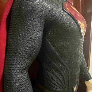 May include: A black and silver Superman costume with a red cape. The suit features a textured pattern and the iconic Superman logo on the chest. The cape is a deep red color.