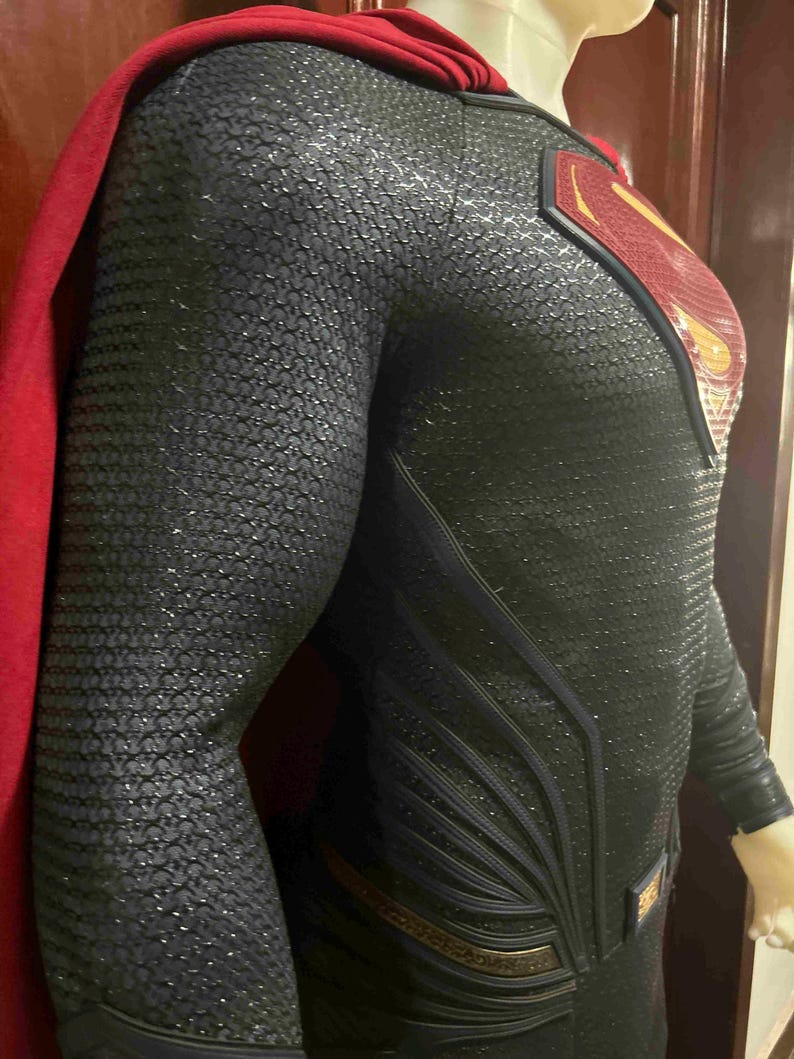 May include: A black and silver Superman costume with a red cape. The suit features a textured pattern and the iconic Superman logo on the chest. The cape is a deep red color.