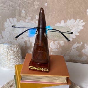 May include: A wooden eyeglass holder with a dark brown finish, holding a pair of rimless glasses. The holder sits atop a stack of colorful books with orange, yellow, and green covers. The base of the holder has a decorative gold design.