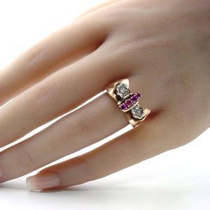 May include: A gold ring featuring a central row of four small round red stones flanked by two white metal floral-shaped settings, each containing a single clear stone. The band is polished gold and has a wide, flat profile across the finger.