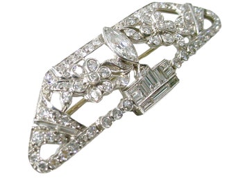 May include: A silver brooch with a large, clear, marquise-shaped diamond in the center. The brooch is decorated with smaller diamonds arranged in a floral pattern.
