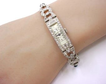 May include: A rectangular silver-toned wristwatch featuring a white dial with black Arabic numerals and dark hands. The watch case and the articulated link bracelet are entirely encrusted with small, clear, sparkling stones throughout.