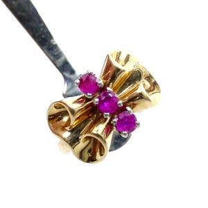 May include: A gold brooch featuring three bright red gemstones. The brooch has a distinctive design with curved, tube-like shapes. The gemstones are set in a silver-coloured metal.