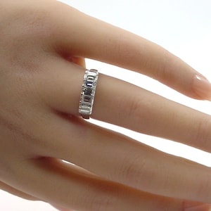May include: A silver-toned band ring featuring a row of five rectangular-cut clear stones set in a channel or bar setting. The ring is crafted from a polished metallic material that reflects light, creating a bright and uniform appearance on the finger.