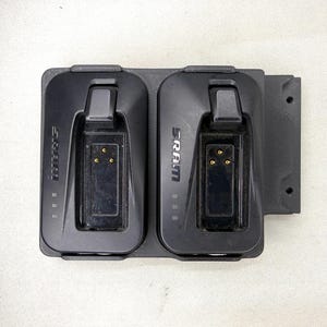 May include: A black dual battery charging dock featuring two slots with the SRAM logo printed on each side. The unit includes integrated locking latches, three gold contact pins per slot, and a textured mounting plate with two circular screw holes.