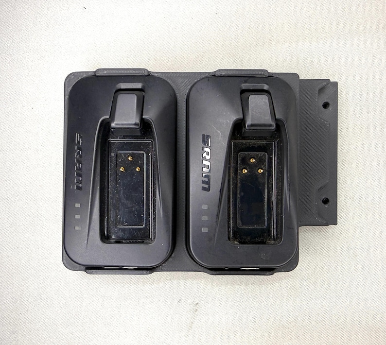 May include: A black dual battery charging dock featuring two slots with the SRAM logo printed on each side. The unit includes integrated locking latches, three gold contact pins per slot, and a textured mounting plate with two circular screw holes.
