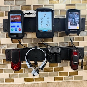 May include: A black wall-mounted storage rack holding three cycling computers, two red taillights, and a pair of white and black bone conduction headphones. The top row features a Garmin, a Wahoo, and a Hammerhead device, with a Wahoo logo plate.