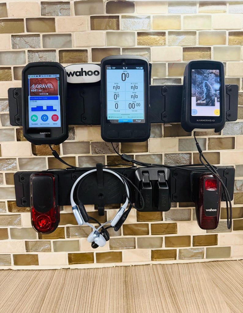May include: A black wall-mounted storage rack holding three cycling computers, two red taillights, and a pair of white and black bone conduction headphones. The top row features a Garmin, a Wahoo, and a Hammerhead device, with a Wahoo logo plate.