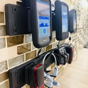 May include: A black wall-mounted storage rack holding several cycling accessories, including Garmin GPS units with screens displaying data, a red rear bicycle light, and a pair of light blue bone conduction headphones.