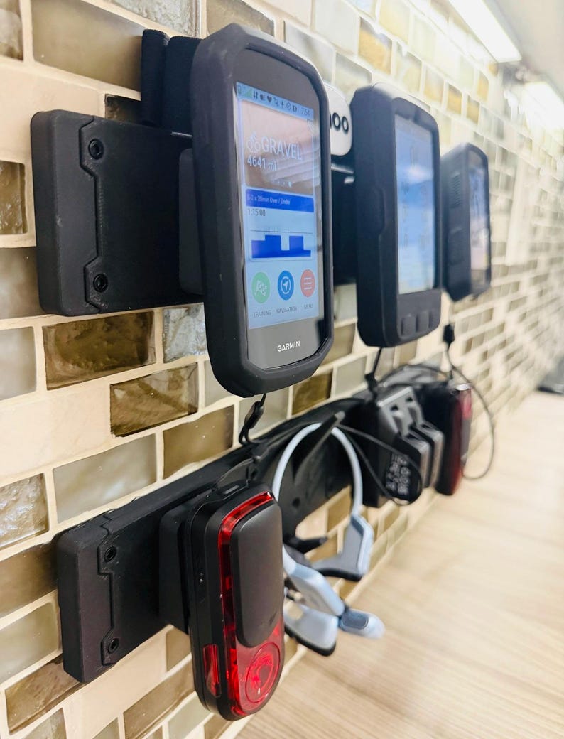 May include: A black wall-mounted storage rack holding several cycling accessories, including Garmin GPS units with screens displaying data, a red rear bicycle light, and a pair of light blue bone conduction headphones.