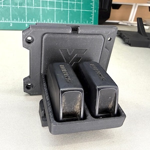 May include: A black 3D-printed plastic wall mount featuring a square backplate with four screw holes and a front-facing cradle holding two black SRAM electronic bicycle derailleur batteries, each clearly labeled with the SRAM brand name.