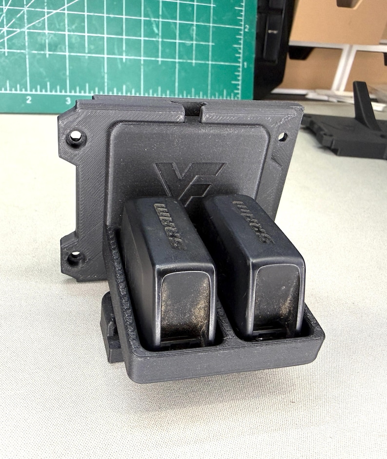 May include: A black 3D-printed plastic wall mount featuring a square backplate with four screw holes and a front-facing cradle holding two black SRAM electronic bicycle derailleur batteries, each clearly labeled with the SRAM brand name.