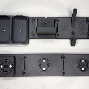 May include: Two black matte 3D printed mounting plates. The top piece features two rectangular storage compartments, a central bracket with the letters VF, and a vertical hook. The bottom piece has three circular locking mounts with screws.