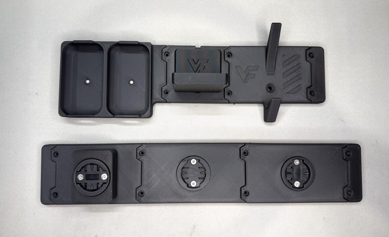 May include: Two black matte 3D printed mounting plates. The top piece features two rectangular storage compartments, a central bracket with the letters VF, and a vertical hook. The bottom piece has three circular locking mounts with screws.