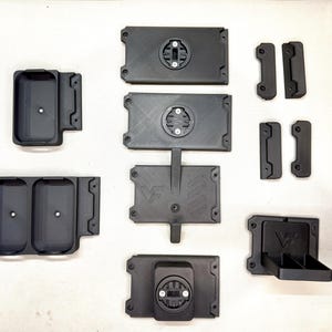 May include: A collection of black 3D-printed plastic components including rectangular mounting plates with circular locking mechanisms, storage trays with single or double compartments, and small brackets, some featuring a VF logo engraving.