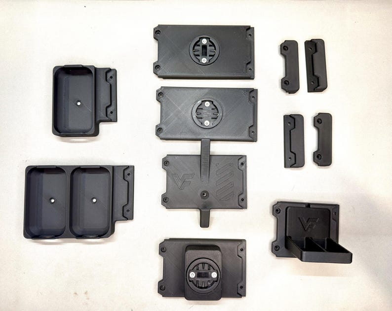 May include: A collection of black 3D-printed plastic components including rectangular mounting plates with circular locking mechanisms, storage trays with single or double compartments, and small brackets, some featuring a VF logo engraving.