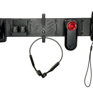 May include: A black wall-mounted organizer rack featuring integrated slots and hooks. It holds two charging docks, a pair of wireless bone conduction headphones, a red rear bicycle light, and a digital cycling computer with a wrist lanyard.