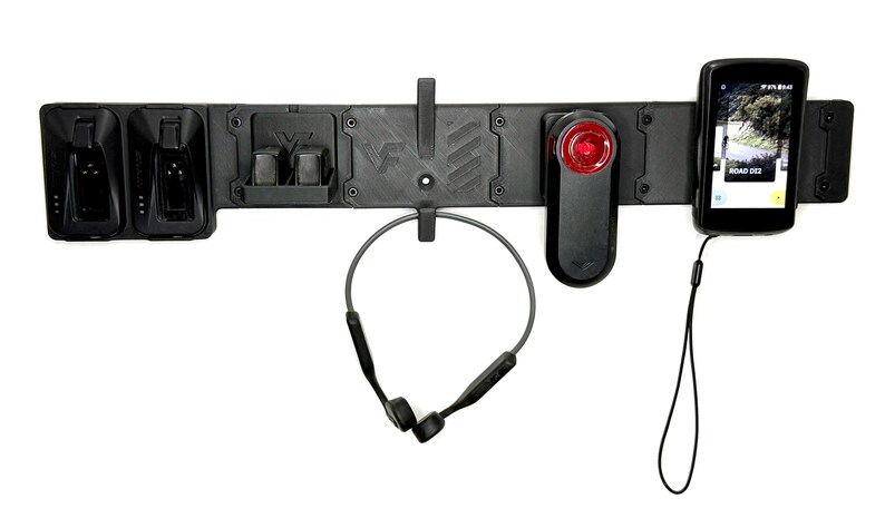 May include: A black wall-mounted organizer rack featuring integrated slots and hooks. It holds two charging docks, a pair of wireless bone conduction headphones, a red rear bicycle light, and a digital cycling computer with a wrist lanyard.