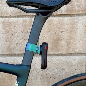 May include: A black bicycle seat with a perforated surface is mounted on a dark gray seat post. A teal and purple clamp holds a red taillight. The bicycle frame is dark teal.