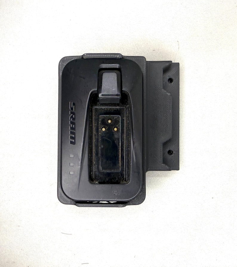 May include: A black SRAM battery charger featuring a rectangular housing with a central recessed port containing three gold-colored electrical contact pins. The unit includes a side mounting bracket with two circular holes and the SRAM logo printed on the front.