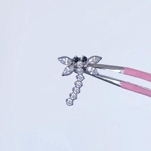 May include: A small, silver-toned dragonfly-shaped jewelry piece. The dragonfly has clear, sparkling accents and black eyes. It is held by pink-tipped tweezers against a white background.