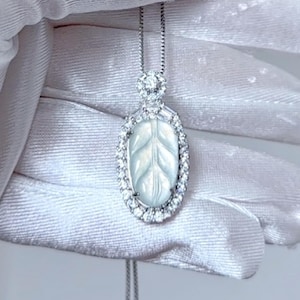 May include: A silver pendant necklace featuring an oval-shaped pendant with a leaf design. The pendant is surrounded by a halo of small, clear gemstones. The necklace chain is delicate and silver.