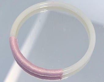 May include: A translucent white bangle bracelet with a section wrapped in pink thread. The bracelet is circular and appears to be made of a smooth, possibly plastic, material. The pink thread adds a decorative element to the accessory.