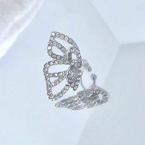 May include: A silver-toned butterfly-shaped clip-on earring. The earring is encrusted with small, clear crystals, creating a sparkling effect. The butterfly wings are open, and the design is detailed and elegant.