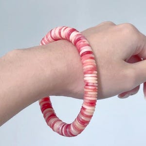 Könnte beinhalten: Ein Armband aus roten und weißen kreisförmigen Perlen. Das Armband wird am Handgelenk getragen und hebt die kontrastierenden Farben und die Textur der Perlen hervor. Das Armband ist ein Schmuckstück.