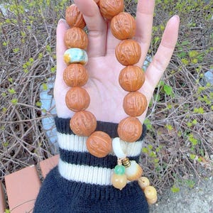 Pode incluir: Uma pulseira de contas com grandes contas redondas marrons, semelhantes a nozes. A pulseira inclui uma conta azul clara e branca, e um berloque com contas verdes e amarelas. A pulseira é exibida em um fundo de folhagem verde.