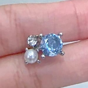 May include: A small cluster earring with a light blue gemstone, a clear gemstone, and a white pearl. The gemstones are set in a silver-coloured metal setting. The earring is a delicate piece of jewellery.