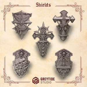 May include: Five gray fantasy shields with intricate designs are displayed. The shields feature various shapes, including crosses and crests, with details like skulls, wings, and decorative elements. The word "Shields" is at the top, and the GreyTide Studio logo is at the bottom.
