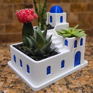 May include: A white planter shaped like a Greek village with blue accents. It features a red cactus, green succulents, and tall green plants. The planter has a blue dome and arched blue windows.