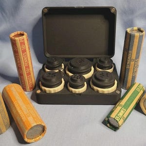 May include: A black coin organizer with various coin rolls and coin wrappers. The rolls are labeled with denominations like "$25 Dollars", "50 Pennies", and "$5 Dimes". The organizer holds coin counting trays.