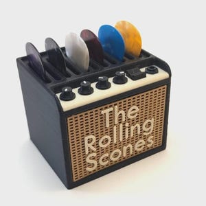 May include: A black and tan guitar pick holder shaped like a vintage amplifier, with the text "The Rolling Scones" on the front. The holder contains several guitar picks in various colors.