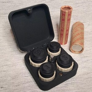 May include: A black coin holder with four compartments for coins. Each compartment has a black lid with a number on it. The holder is open and the lids are off. There are two rolls of coins next to the holder. The rolls are labeled "Pennies 50 Pennies 50 Pennies 50 Pennies 50 Pennies 50 Pennies".