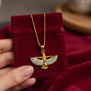 May include: A gold-colored pendant necklace featuring an Egyptian-inspired design with a figure and outstretched wings, accented with light blue and white enamel. The necklace is displayed on a red velvet pouch.